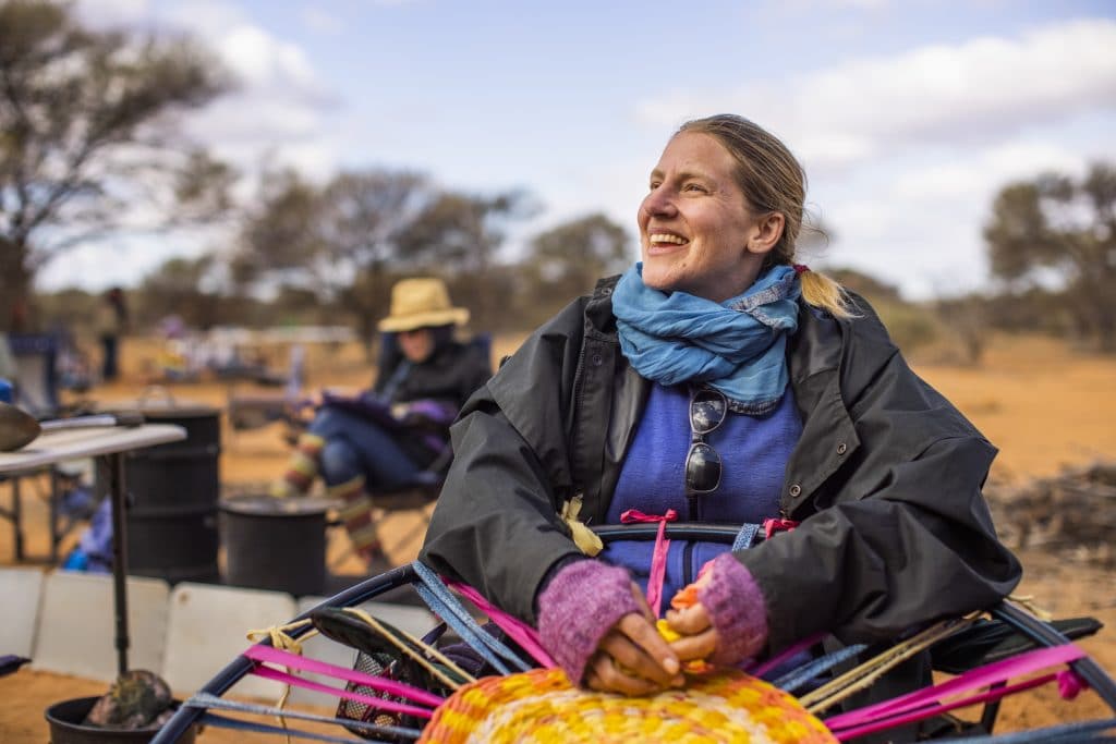 artist Lucy Ridsdale smiling and taking a break from weaving a rag rug sitting in the outback