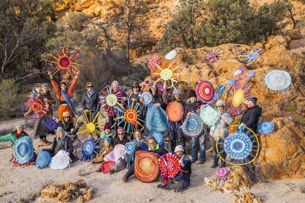 tens of handmade colourful rag rugs laid out on the rocks and held up by heaps of people in the outback