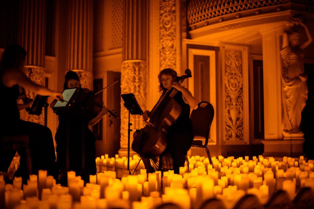 A woman plays the cello surrounded by flameless candles in an ornate venue.
