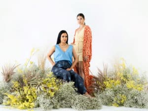 Two First Nation women wearing Indigenous designer clothing by KIRRIKIN, sitting amongst native flowers and plants