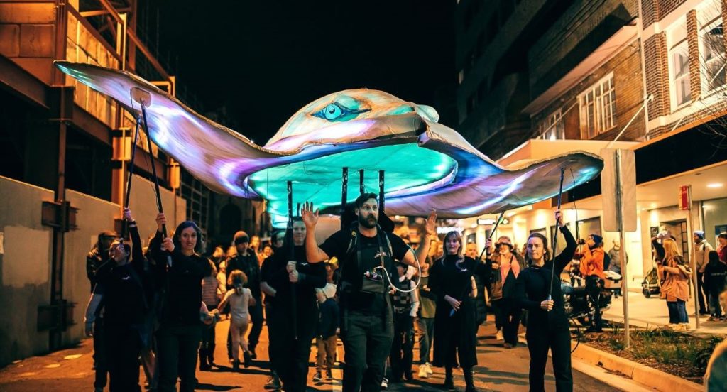 Crowd of people walk beneath giant illuminated stingray puppet